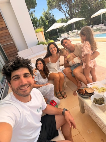 Un hombre con barba sonríe a cámara tomando una selfie grupal con cuatro adultos y dos niños sentados en un patio junto a una piscina al aire libre