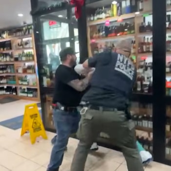 Un oficial del NYPD con uniforme forcejea con un hombre vestido con camiseta negra y jeans dentro de una tienda. Al fondo se ven estantes con botellas y un letrero amarillo de 'piso mojado'