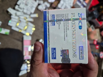 Una mano sostiene una caja blanca y azul de fluoximesterona de 10 mg y 100 comprimidos. Se aprecian otras cajas blancas en el fondo difuso