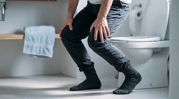 Persona levantándose de un inodoro, con las manos apoyadas en los muslos. Se ven sus piernas, calcetines oscuros y el inodoro. Hay una toalla y papel higiénico.