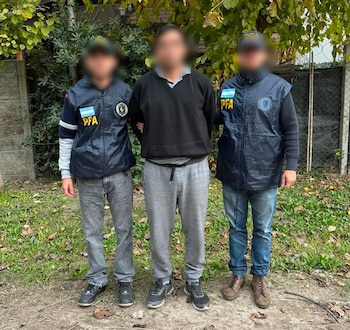 Dos agentes de la Policía Federal Argentina con chalecos azules flanquean a un individuo en ropa civil, de pie sobre tierra con árboles y una valla detrás
