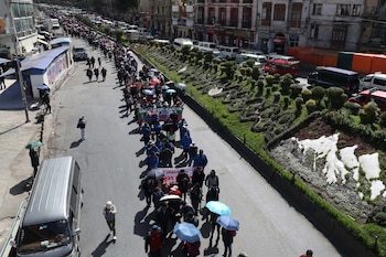 Vista aérea de una marcha de personas en una calle urbana. Edificios y vehículos a los lados. Hay una mediana con plantas y un mosaico blanco. Algunas personas llevan sombrillas