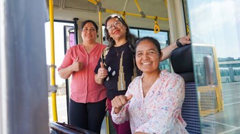 Tres mujeres adultas sonriendo, dos con pulgares arriba, posan dentro de un autobús; al fondo, otros autobuses y cielo despejado