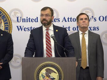 Un hombre con traje habla ante un podio con el sello del Departamento de Justicia junto a dos hombres y un cartel de la fiscalía
