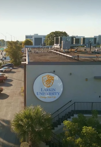 Primer plano de un logo circular blanco y azul de Larkin University en un edificio gris, con una palmera Sabal en la esquina inferior y edificios en la distancia