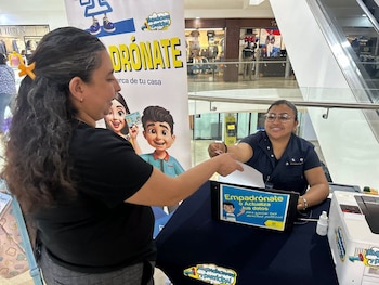 Una mujer entrega un documento a una atendiente sentada tras un escritorio con carteles de empadronamiento electoral dentro de un centro comercial