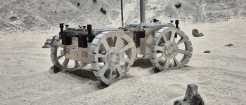 Un rover de metal plateado con grandes ruedas de radios y tacos rectangulares se asienta sobre arena clara, con un fondo rocoso que simula un paisaje extraterrestre