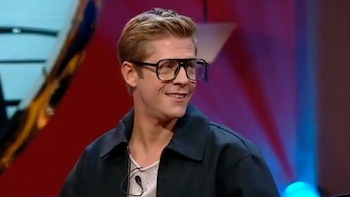 Hombre de cabello rubio y gafas de montura gruesa, con chaqueta oscura y camiseta clara, sonríe ligeramente mirando a la derecha. Fondo de luces de estudio rojas y azules