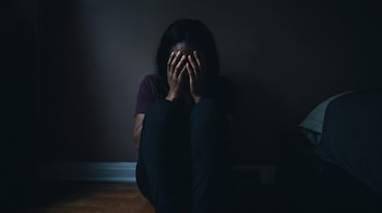 Una mujer sentada en el suelo de madera, con piel morena, cubre su rostro con ambas manos. Viste camiseta y pantalón, en un cuarto oscuro con una cama al lado.