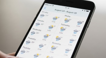 Una mano sostiene un teléfono móvil con una aplicación del clima abierta que muestra la semana del 24 al 30 de agosto, iconos de clima y temperaturas.