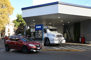 Vista de una estación de servicio YPF con cartel de precios de combustible, un SUV rojo en movimiento y un camión blanco estacionado bajo la marquesina en un día soleado