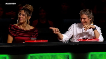 Una mujer con un vestido rojo escotado y un hombre con camisa blanca sentados en un panel. El hombre sonríe y señala; la mujer tiene una leve sonrisa