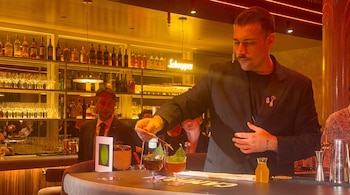 Un hombre con bigote decora un cóctel con pinzas. En la barra hay otro cóctel y un vaso. Al fondo se ven botellas, copas y un letrero de neón.