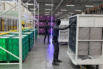 Dos robots humanoides, estanterías con cajas verdes y moradas, carro de almacenamiento y pasillo de almacén industrial