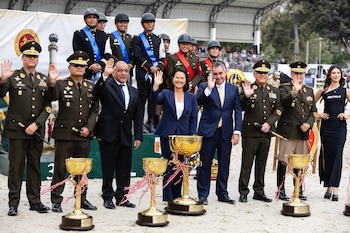 Un grupo de militares, civiles y jinetes, entre ellos una mujer, saludan al frente de trofeos dorados y un cartel. Todos están en una pista de arena