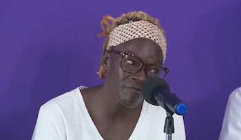 Una persona con rastas rubias, diadema de encaje beige y gafas marrones habla frente a un micrófono oscuro sobre un fondo morado liso