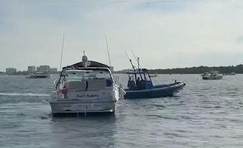 Una lancha blanca en primer plano con el nombre 'Naoli Nakora', junto a un bote azul. Más embarcaciones y edificios costeros se ven en la distancia