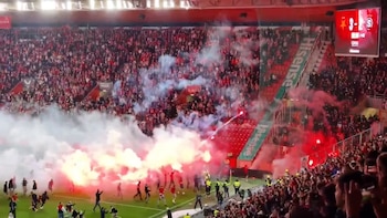 Vista aérea de un estadio de fútbol lleno de gente, con el campo invadido por aficionados y humo blanco y rojo de bengalas encendidas en primer plano