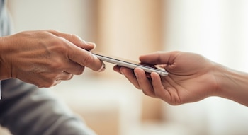 Primer plano de una mano arrugada entregando un teléfono inteligente plateado a una mano más joven y suave, con un fondo difuminado.