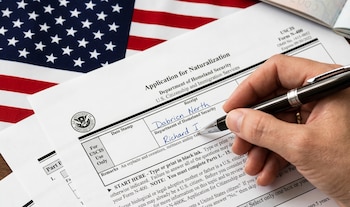 Primer plano de una mano escribiendo con un bolígrafo sobre el Formulario N-400 de naturalización de USCIS, con la bandera de Estados Unidos desenfocada detrás