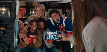 Cuatro jóvenes sonríen y gritan de emoción dentro de una furgoneta, mientras una persona con el pelo largo les toma una foto con un teléfono móvil