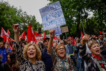 Varias mujeres en primer plano gritan y alzan llaves en una calle con árboles, acompañadas por personas con banderas rojas y carteles