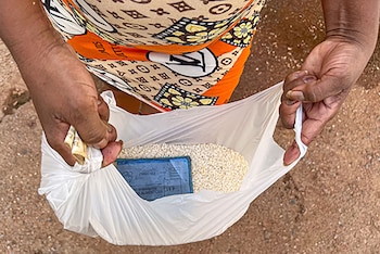 Vista cenital de dos manos sujetando una bolsa plástica transparente con arroz y una libreta de racionamiento azul sobre un fondo de tierra