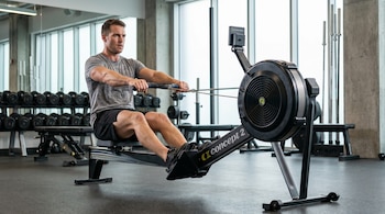 Hombre caucásico concentrado en una máquina de remo Concept 2, vistiendo camiseta gris y pantalones cortos negros, en un gimnasio con ventanales grandes y estantes de pesas.