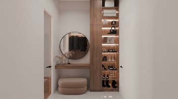 Vista frontal de un vestidor moderno con paredes blancas, un armario de madera, estantes iluminados con zapatos y bolsos, un tocador flotante, espejo redondo y una otomana beige.