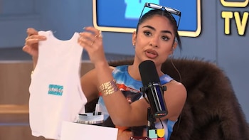 Mujer sostiene remera blanca sin mangas con diseño brillante. Viste camiseta estampada, lentes de sol en cabeza y brazaletes dorados. Micrófono y fondo azul