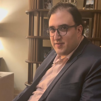 Un hombre con camisa rosa, saco oscuro y gafas de marco negro mira hacia adelante, con un estante de madera y fotografías al fondo