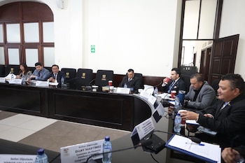 Diputados sentados en una mesa curva con micrófonos, documentos, botellas de agua e identificadores de nombres en una sala de reuniones
