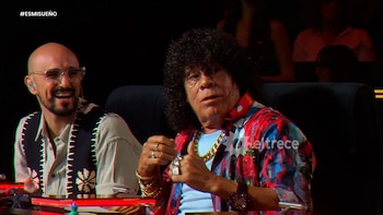 Dos hombres sentados en un set oscuro. Un hombre calvo con gafas sonríe a la izquierda. A la derecha, un hombre de pelo rizado, camisa colorida y joyas de oro hace un gesto con la mano