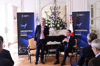 Luis Caputo habla de pie con un micrófono junto a Diego Santilli, quien permanece sentado entre carteles promocionales y banderas nacionales
