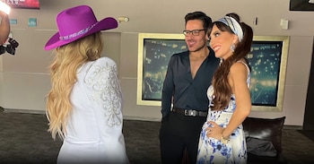 Tres personas en un interior: una mujer de espaldas con sombrero morado, un hombre sonriente con gafas, y una mujer de perfil con vestido floral y pañuelo