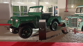 Un vehículo de color verde y una camioneta clásica turquesa aparcados sobre una alfombra roja junto a un pedestal negro