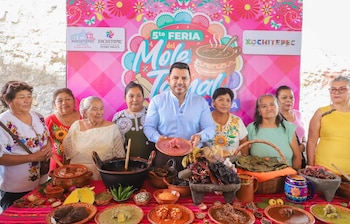Un hombre y ocho mujeres posan frente a una mesa con platos de mole, tamales, chiles y especias. Detrás, un cartel de la Feria del Mole y Tamal