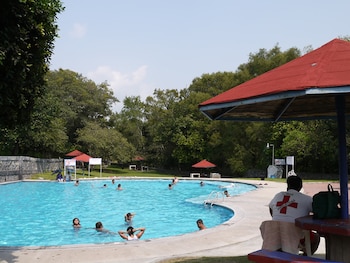 Piscina exterior con bañistas. Un salvavidas bajo una estructura de techo rojo. Vegetación densa rodea la piscina. Varias sombrillas de techo rojo