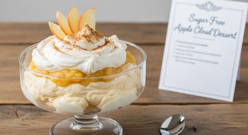 Copa de cristal con postre de manzana sin azúcar, cubierto con crema batida, canela y rodajas de manzana sobre mesa de madera. Tarjeta de receta y cuchara a la derecha.