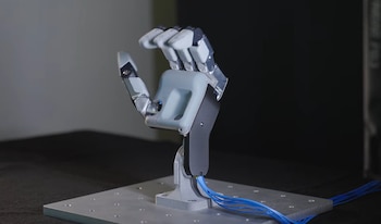 Mano mecánica articulada de color gris y blanco con cables azules, montada sobre una placa metálica con orificios
