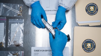 Manos con guantes azules manipulan un arma de fuego plateada sobre mesa de laboratorio. Se observan hisopos, proyectiles y documentos con logo de Fiscalía de Sonora.
