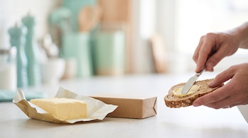 Una barra de mantequilla sobre papel, una caja de cartón y dos manos que untan mantequilla en una rebanada de pan con un cuchillo.