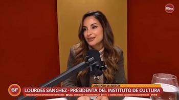 Una mujer con cabello castaño largo y un blazer beige habla frente a un micrófono en un estudio con fondo rojo y amarillo. Se ve un vaso de agua
