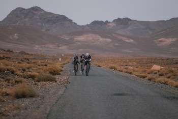 Tres ciclistas con cascos y anteojos de sol circulan en bicicleta por una carretera en medio de un paisaje árido con montañas