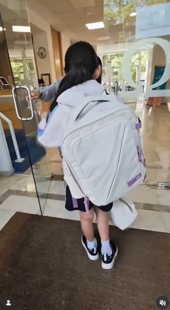 Una niña de espaldas, con una mochila grande blanca y uniforme escolar, abre una puerta de cristal para entrar a un edificio moderno con pisos de baldosas