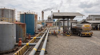 Instalaciones industriales con varios tanques de almacenamiento, tuberías, edificios y un camión cisterna de combustible. Humo de chimeneas al fondo.