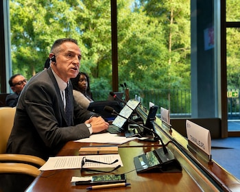 Hombre con traje oscuro y auriculares hablando en una mesa de conferencias con micrófonos, pantallas planas, papeles y gafas, frente a una ventana con árboles verdes