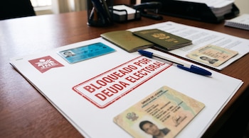 Documentos de identidad, un pasaporte, un bolígrafo azul y un papel marcado con el sello Bloqueado por deuda electoral sobre un escritorio.