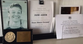 Una foto enmarcada de una mujer sonriente, un certificado del Salón de la Fama de la Natación, una placa conmemorativa y una invitación real