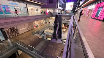 Vista aérea dentro de un centro comercial moderno de varios niveles con tiendas, escaleras mecánicas y personas caminando, con iluminación rosa y púrpura